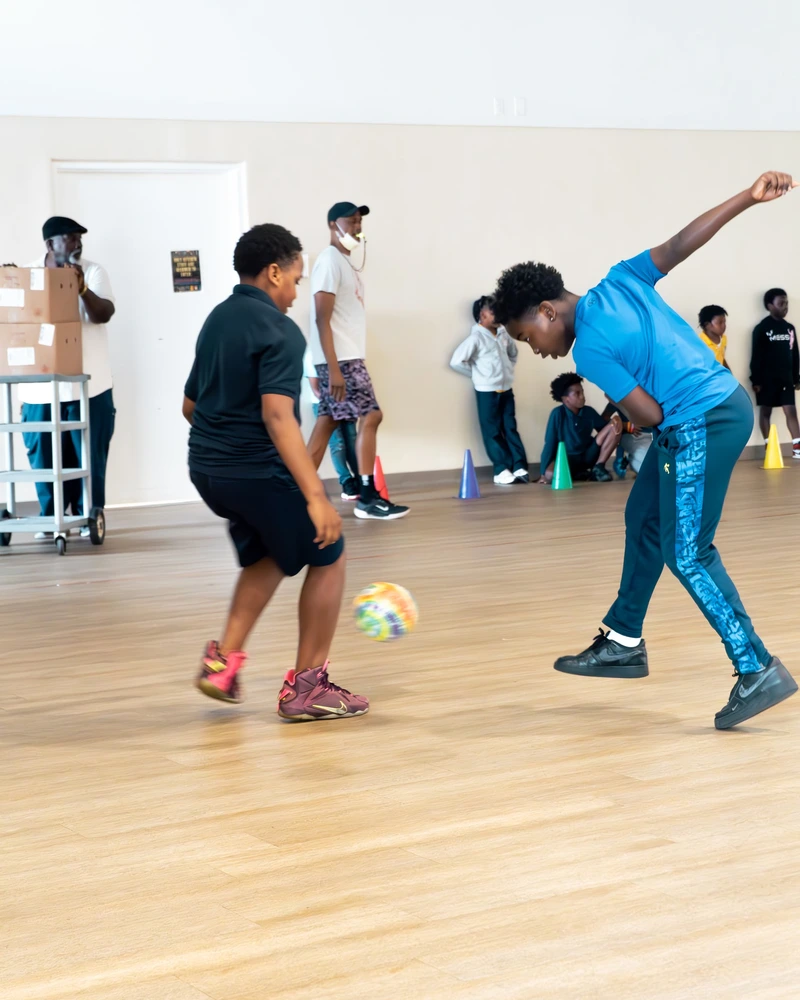 Two children engaged in a playful game, one kicking a colorful ball while others watch from the sidelines in a spacious indoor area.