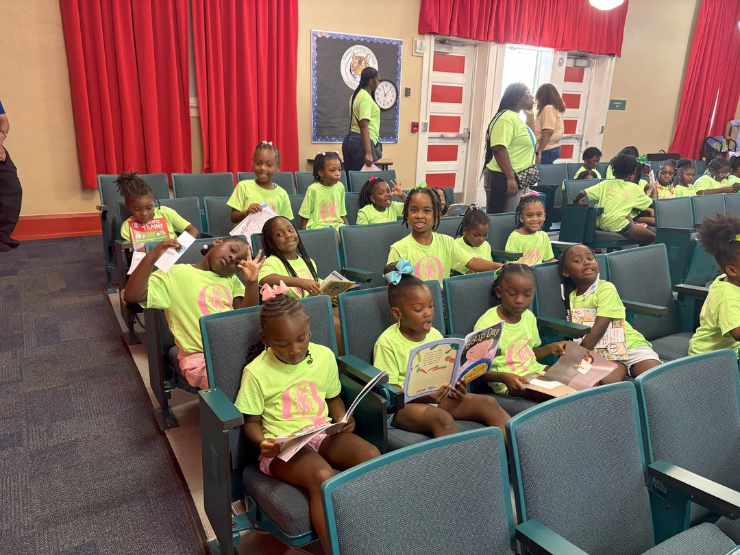 A group of children in bright neon green shirts sit in rows, reading books in a colorful room with red drapes and educational posters.